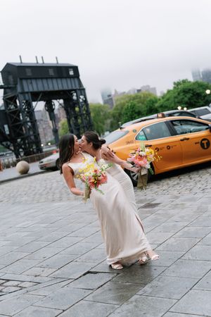Two brides share a romantic kiss on cobblestone streets of NYC, holding colorful bouquets with a yellow taxi in the background.
