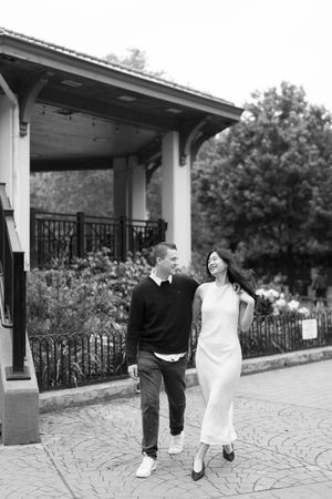 Black and white photo of a smiling couple walking together outdoors near a gazebo surrounded by lush greenery.