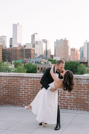 Bride and groom share a romantic dip kiss on a rooftop with a stunning city skyline backdrop at golden hour.
