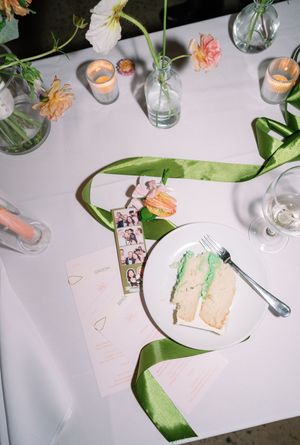 Elegant wedding table setting with green ribbon, cake slice, photo strip, floral arrangements, and candles on white linen.