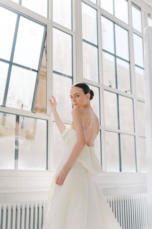 Elegant bride in a strapless white backless wedding gown standing by large windows in a bright airy room.