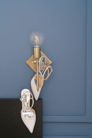 White bridal heels and gold jewelry displayed against a blue wall with a brass globe wall sconce light fixture.