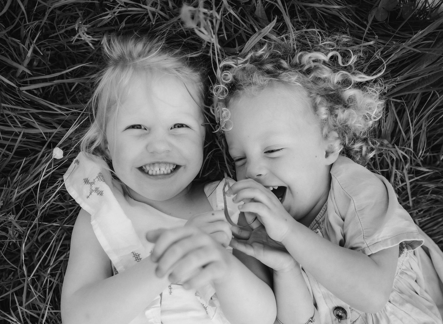 Two young girls laughing and lying in grass, black and white photo capturing joyful childhood. During a family location photoshoot in Sidcup
