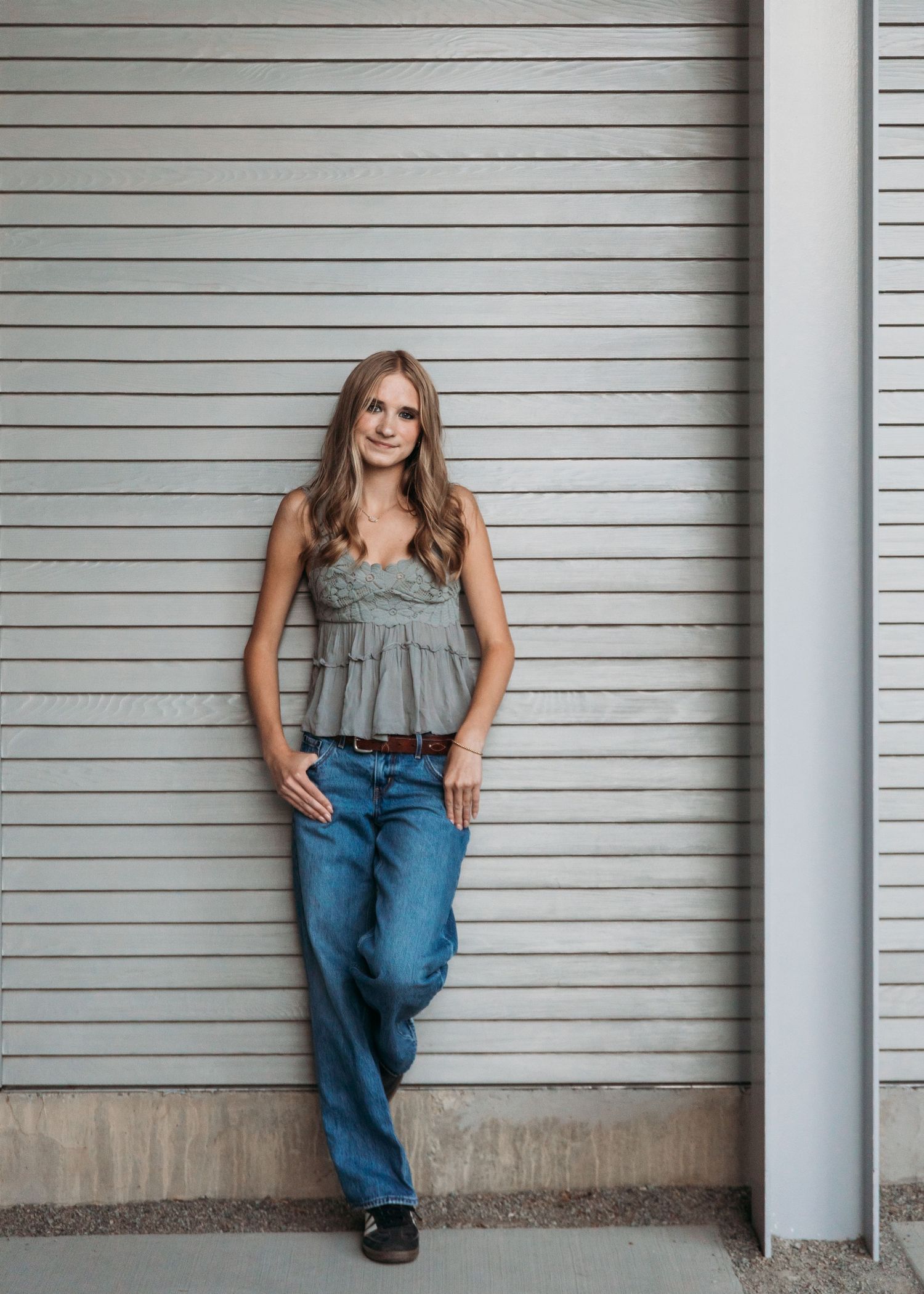 Young woman in gray ruffled tank top and jeans leaning against a corrugated metal wall, smiling confidently.
