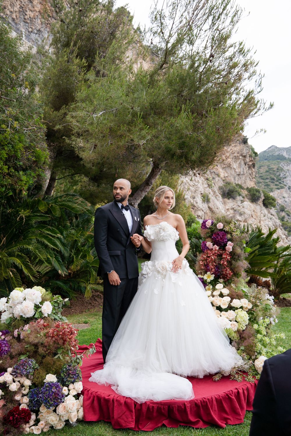 Bride and groom stand on red platform surrounded by flowers at outdoor wedding ceremony with mountain backdrop.