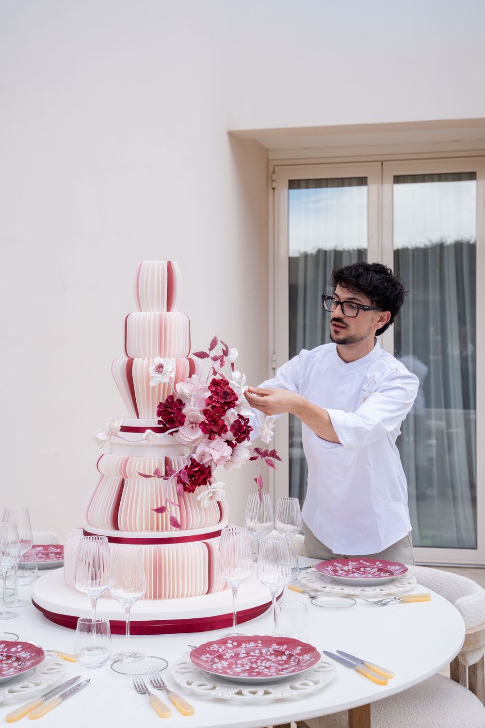 A pastry chef in white uniform decorates an elegant multi-tiered pink wedding cake with red flowers on a table.