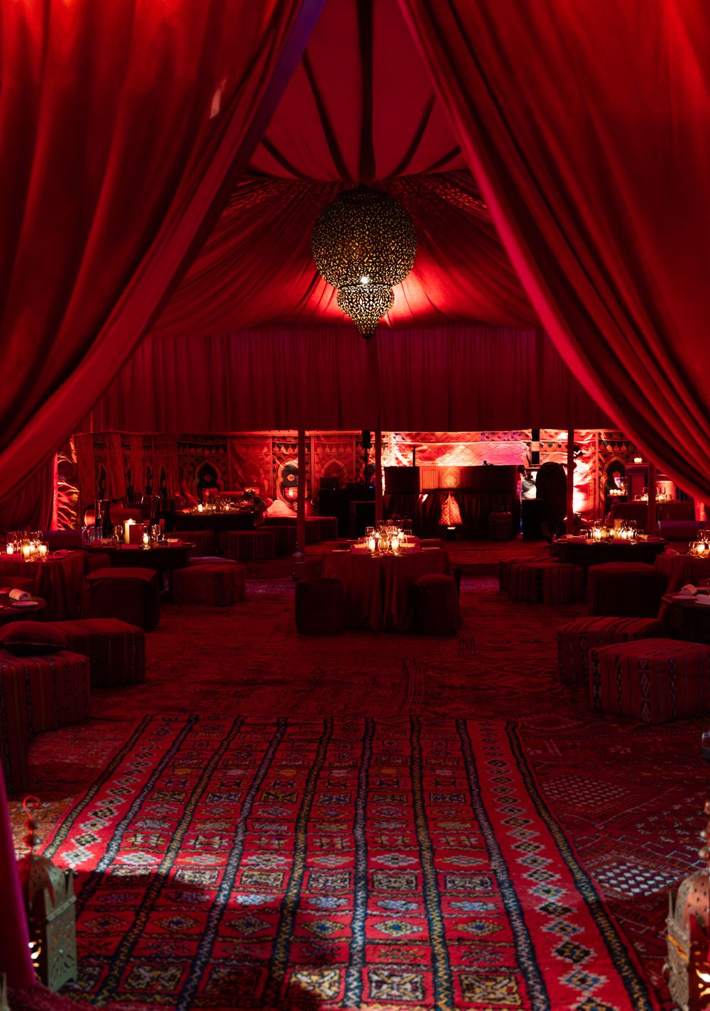 Luxurious Moroccan-themed event tent with red draping, ornate chandelier, patterned rugs, and ambient candlelit lounge seating.