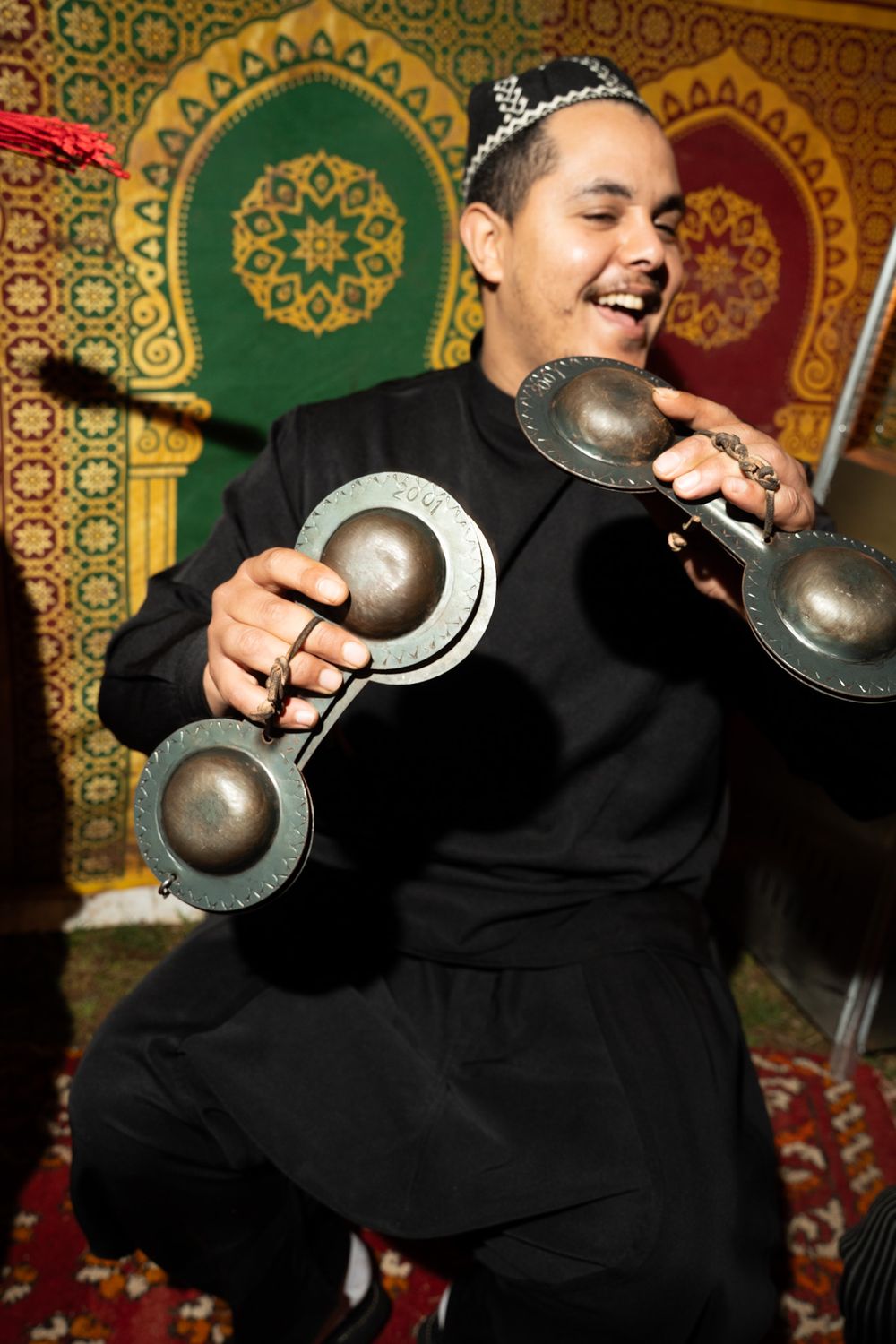 Smiling Moroccan musician playing traditional metal krakeb castanets in black attire with ornate mosaic backdrop.