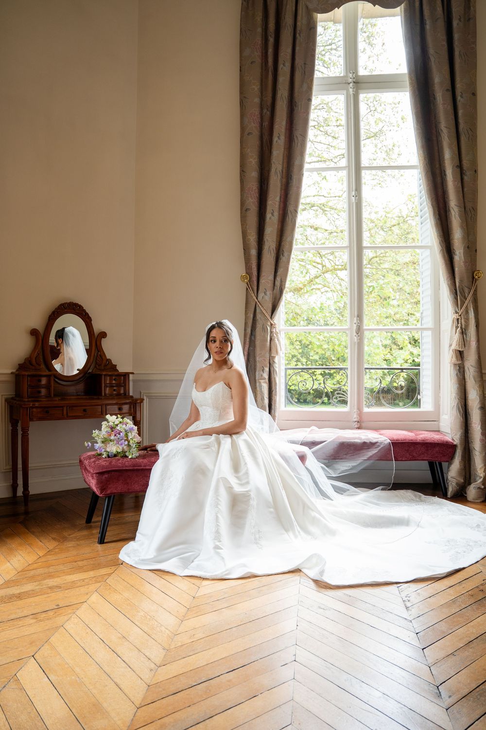 Bride in white gown and veil sits by sunlit window in elegant room with herringbone floors and antique vanity.