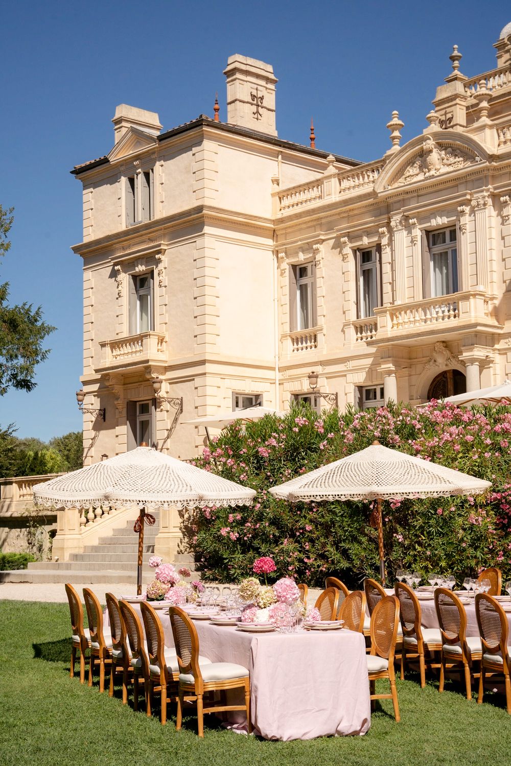 Elegant outdoor wedding reception setup on manicured lawn before grand French-style mansion with pink floral arrangements.
