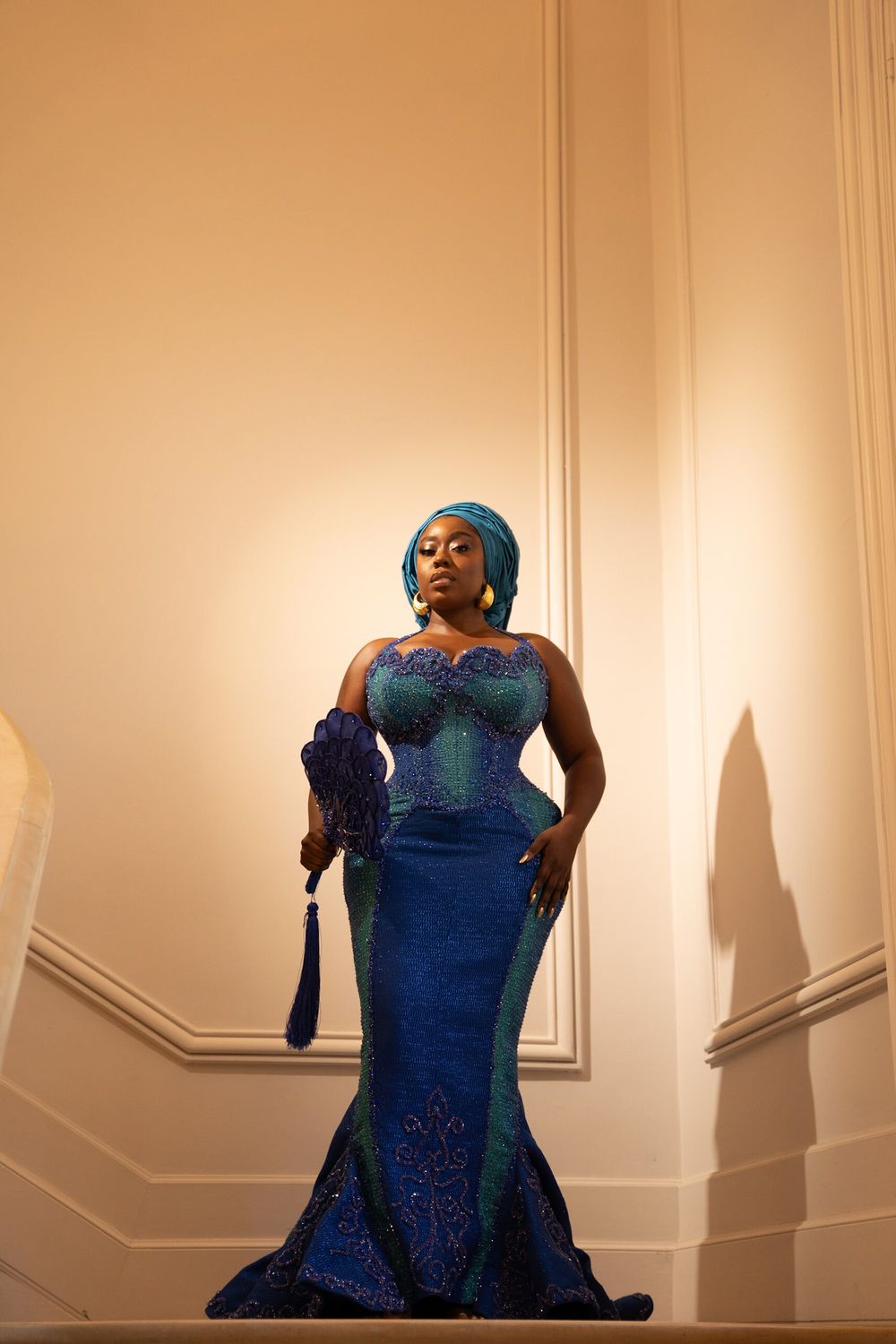Woman in elegant blue African lace gown and teal headwrap poses gracefully on stairs in warm lighting.