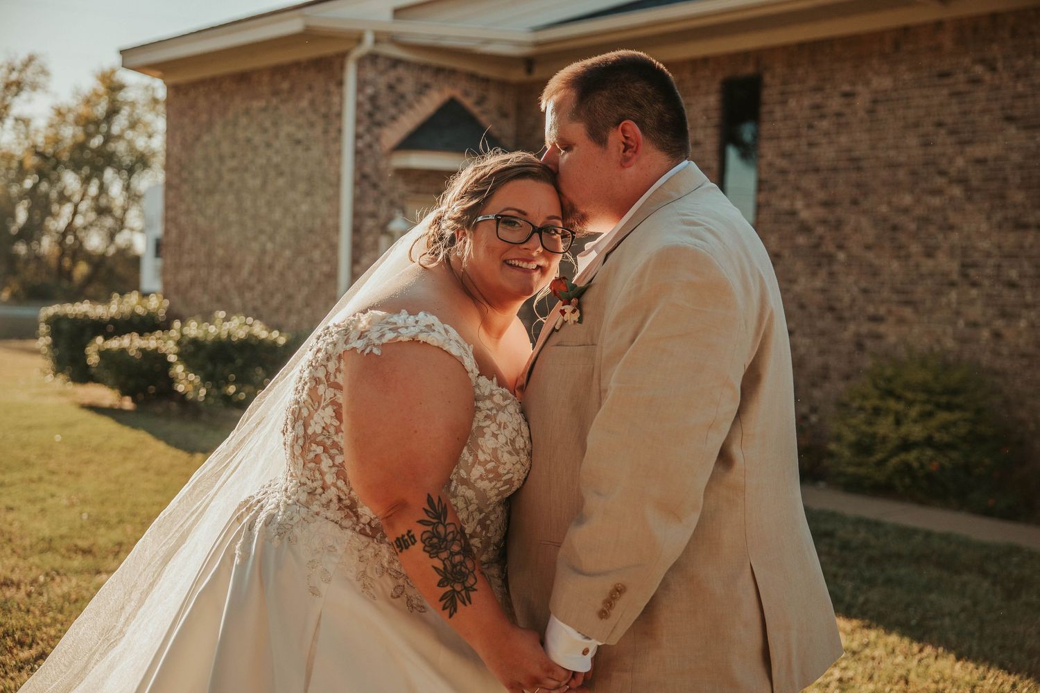 A bride and groom share a tender moment outside a brick church, with the groom kissing the bride's cheek on their wedding day.