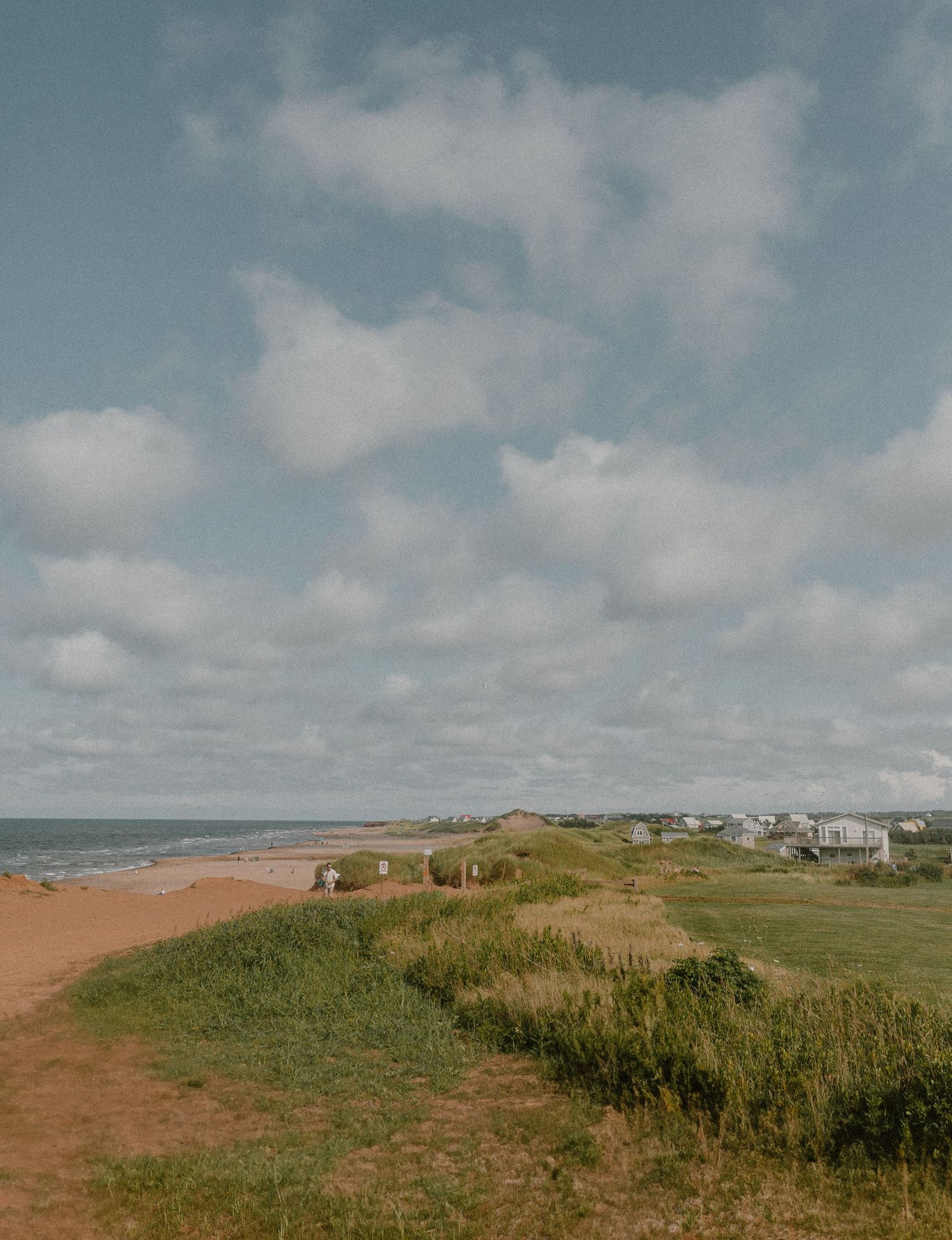 Coastal beach PEI landscape with green dunes, sandy path, ocean waves, and seaside homes under a partly cloudy sky.
