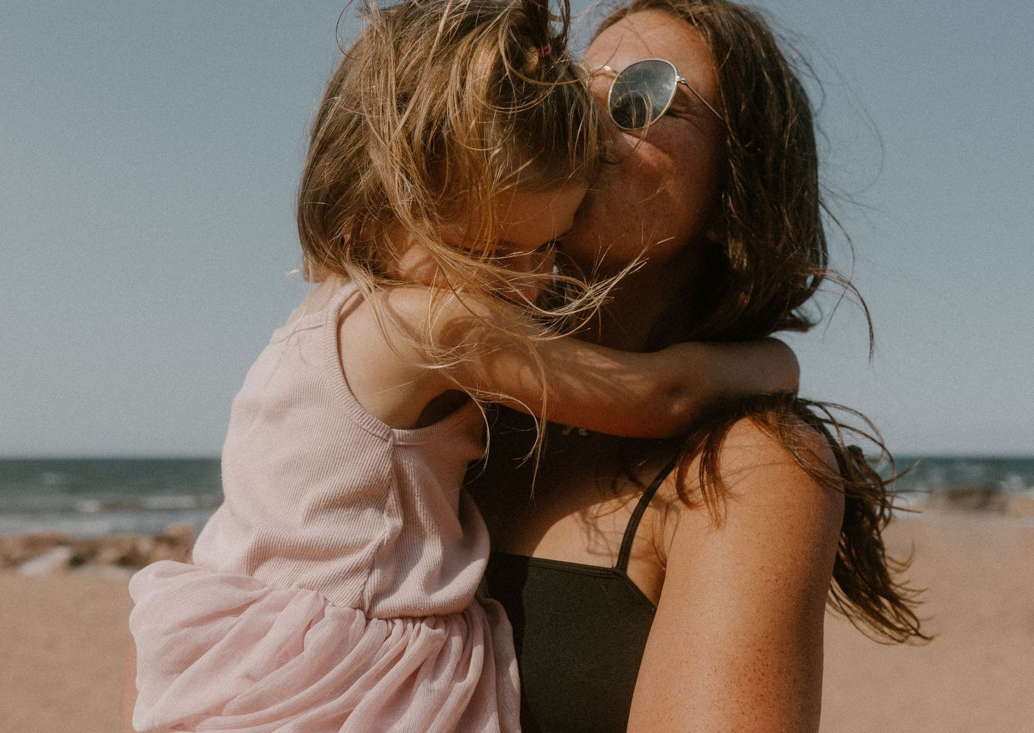 Mother holding and hugging young daughter on sunny beach with ocean in background.