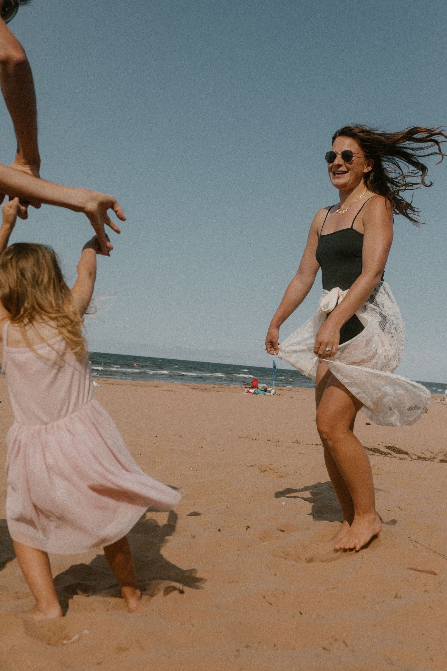 Woman in black swimsuit dancing barefoot on sandy PEI beach with young girl in pink dress.