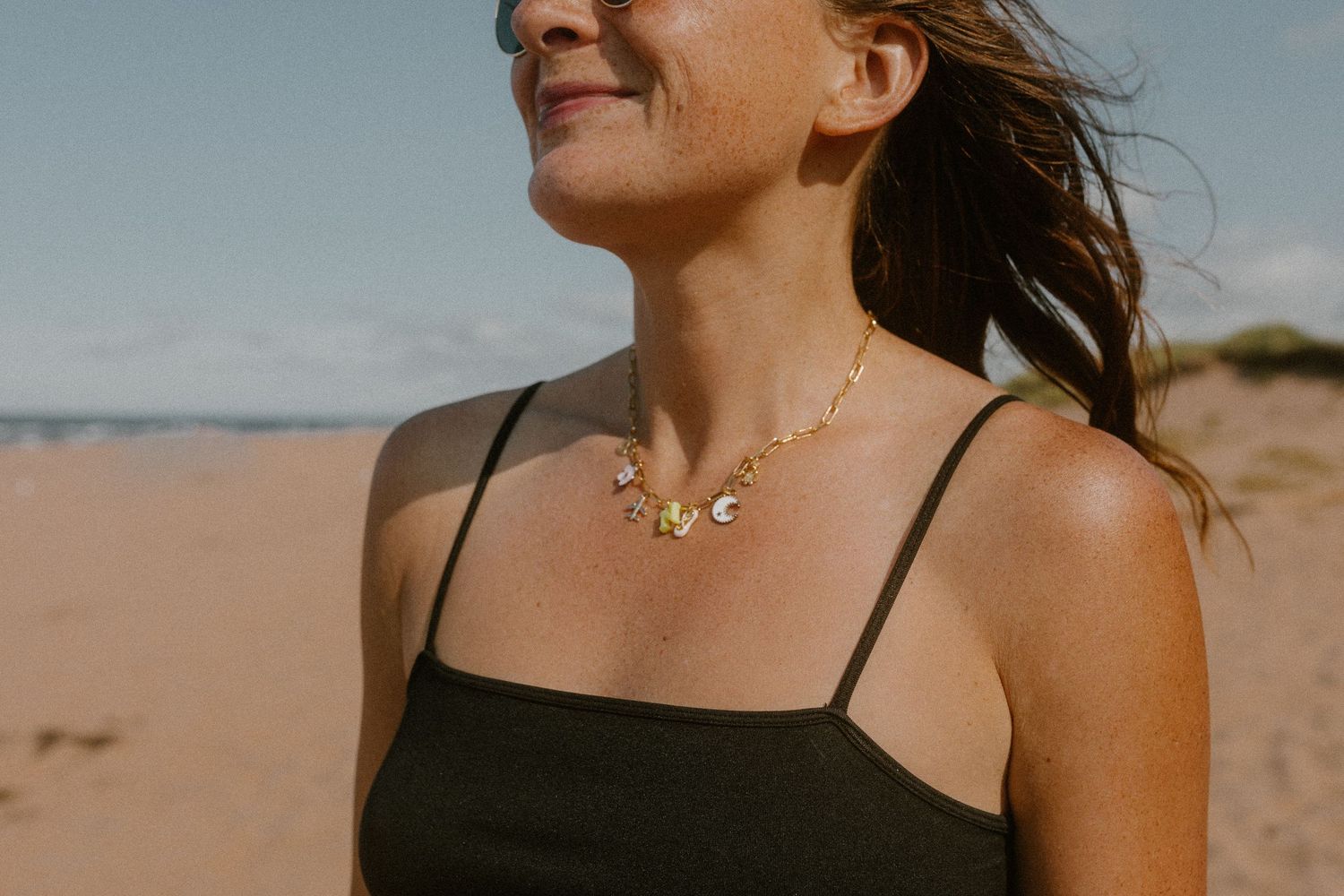 Woman wearing a gold chain necklace with charms on a sunny PEI beach, smiling in a black tank top.