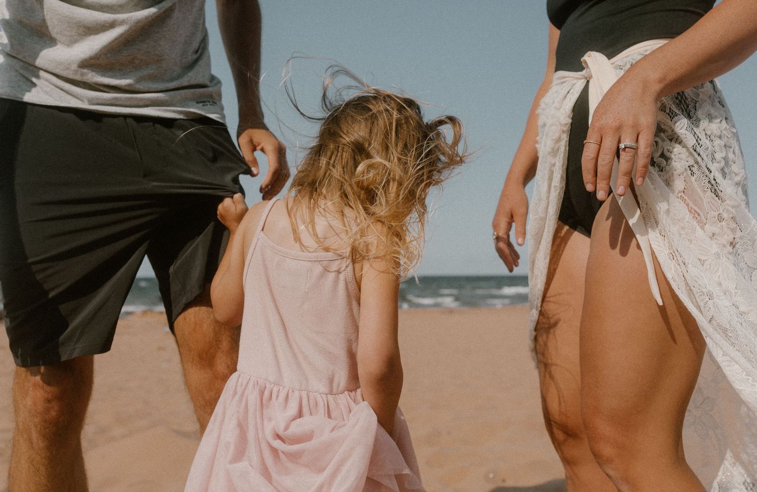 Family with young girl in pink dress walking on sandy PEI beach, parents in casual summer attire near ocean.