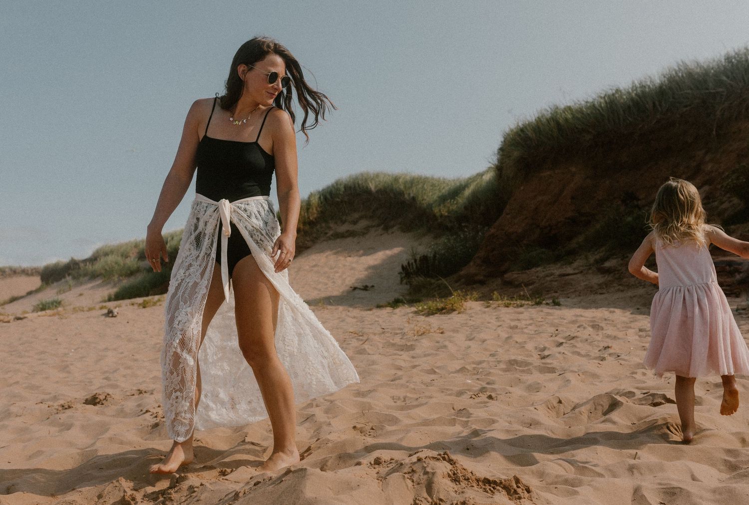 Stylish pregnant woman in black swimsuit and lace wrap walks on sandy PEI beach being chased by young daughter.
