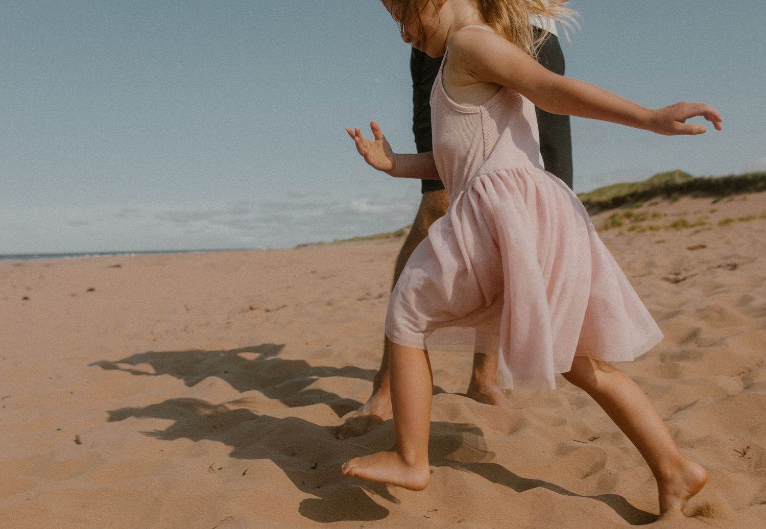 Two barefoot people running playfully on a sandy PEI beach, casting shadows on golden sand with dunes in background.