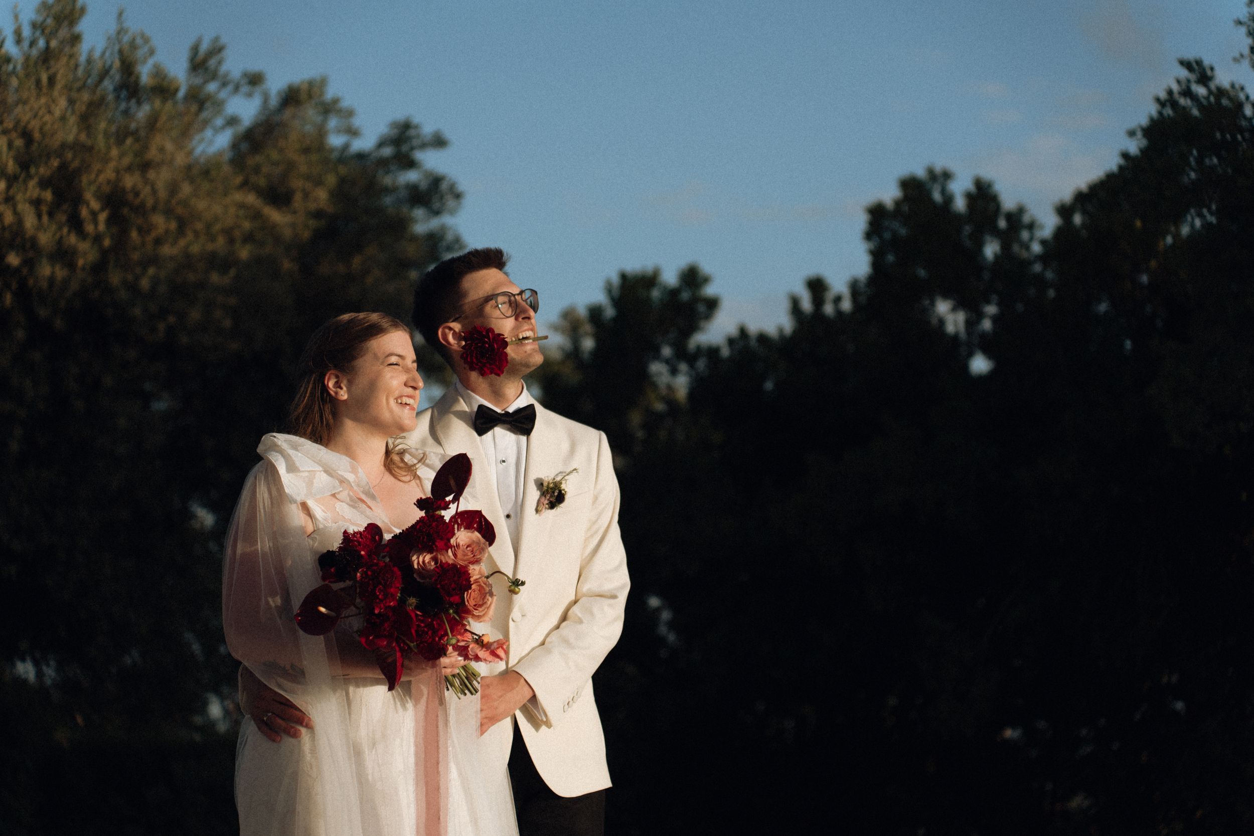 Bride and groom in elegant wedding attire stand together outdoors, smiling, with lush green trees in golden sunlight.