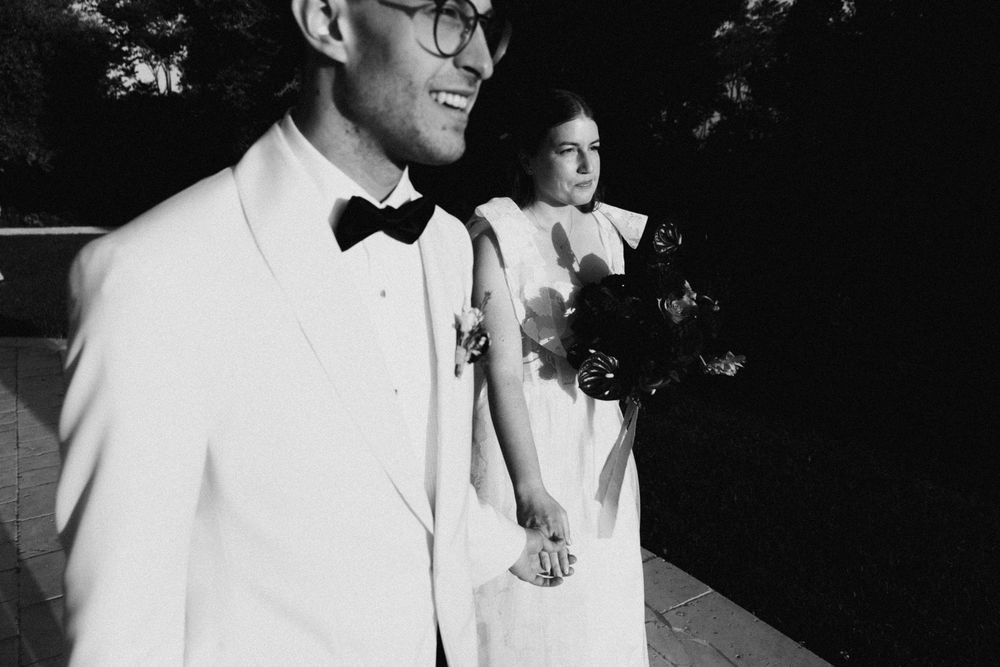 Black and white wedding photo of smiling groom in white tuxedo and bow tie with bride holding bouquet.