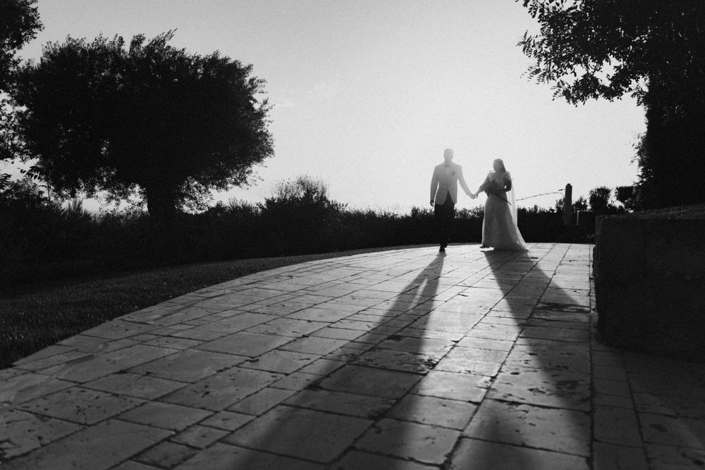 Black and white silhouette of bride and groom walking hand in hand on stone pathway at sunset.
