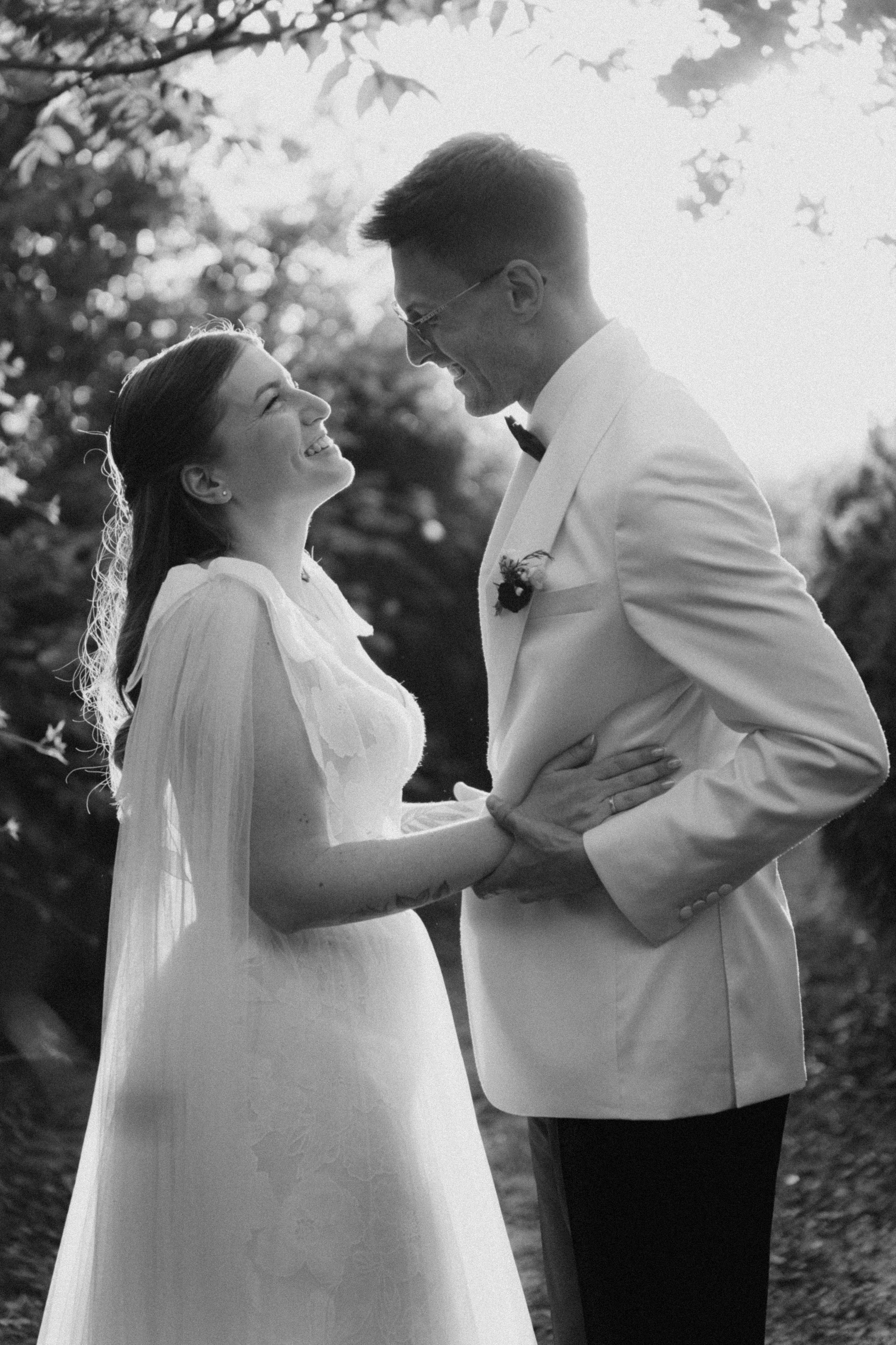 A bride and groom share a joyful moment in black and white, smiling at each other outdoors.