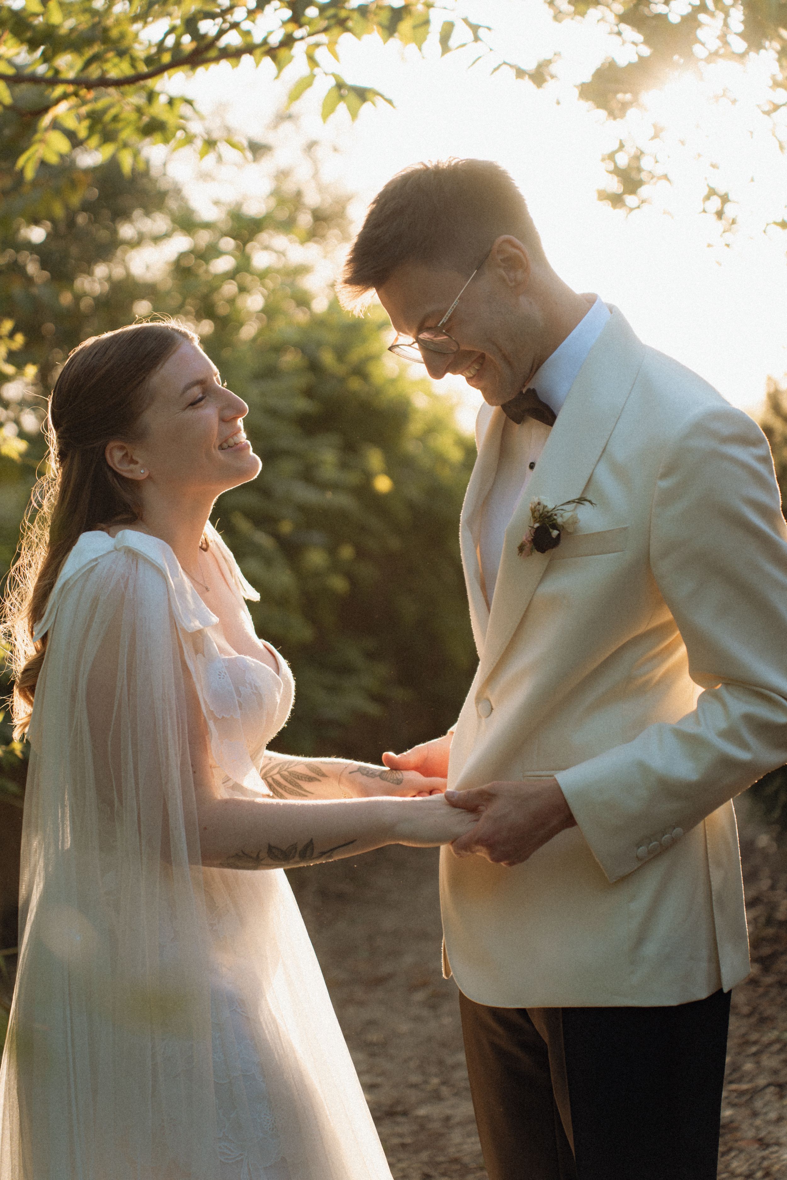 Bride and groom holding hands outdoors in golden sunlight, wearing white wedding attire among lush green trees.