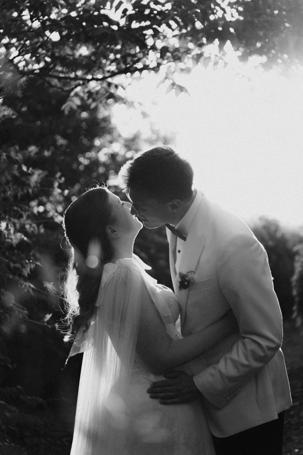 Black and white photo of a bride and groom sharing a romantic kiss outdoors, backlit by soft sunlight.