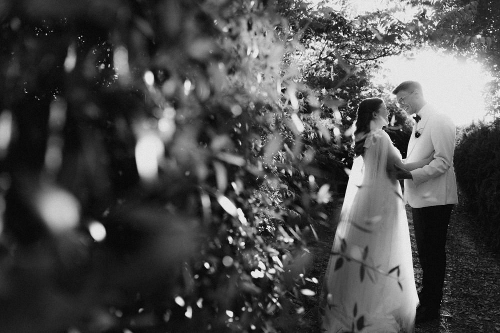 Black and white wedding photo of bride and groom sharing a romantic moment surrounded by lush garden greenery.