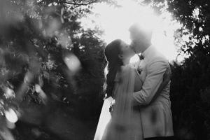 Black and white photo of a couple sharing a kiss outdoors, surrounded by lush trees with soft bokeh light effects.