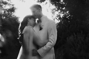 Black and white wedding photo of couple embracing outdoors, bride in veil and groom in light suit, surrounded by lush trees.