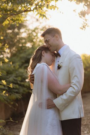 A bride and groom share a tender embrace outdoors during golden hour, with the groom in a white tuxedo kissing her forehead.
