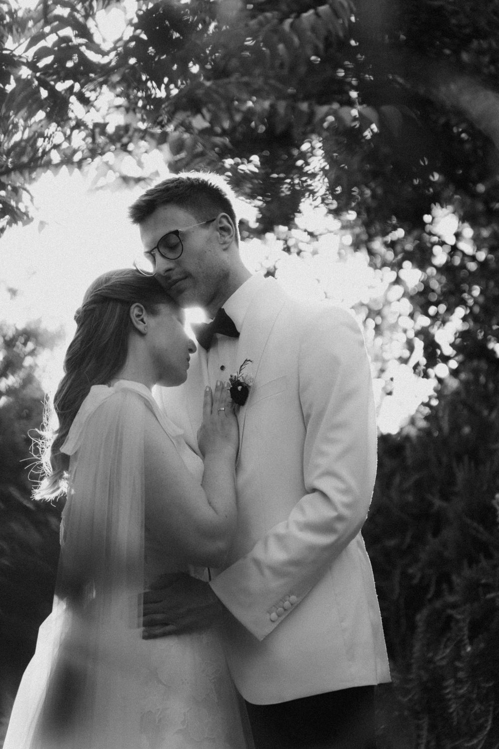 Black and white wedding photo of bride and groom embracing outdoors under trees in elegant attire.
