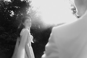Black and white wedding photo of bride in flowing gown glancing back at groom in sunlit outdoor setting.