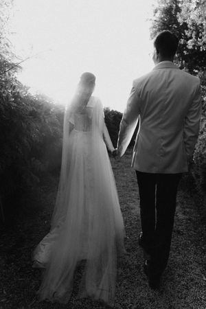 Black and white photo of bride and groom holding hands walking through garden path toward bright light.