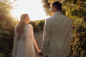 Couple in wedding attire holding hands at golden hour sunset surrounded by lush greenery.