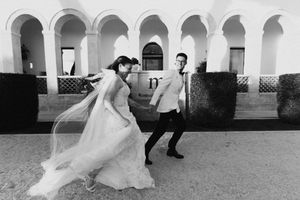 Black and white photo of a bride in flowing gown and groom in suit walking outside arched building.