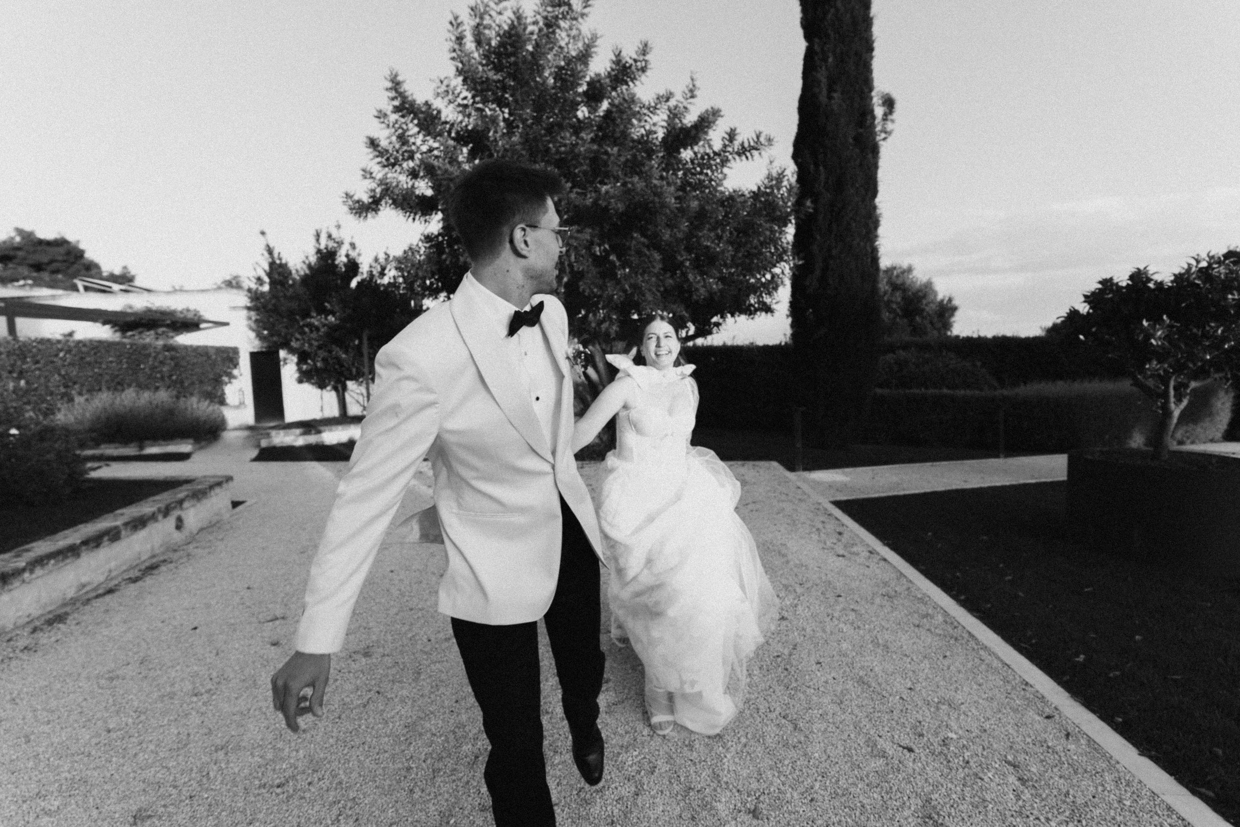 Black and white photo of a bride and groom walking hand in hand along a garden path on their wedding day.
