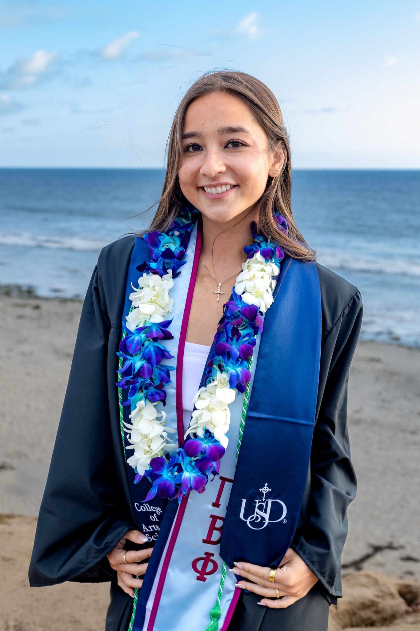 San Diego graduation portrait photography of a female USD graduate posing on the beach in her cap, gown, and floral leis.