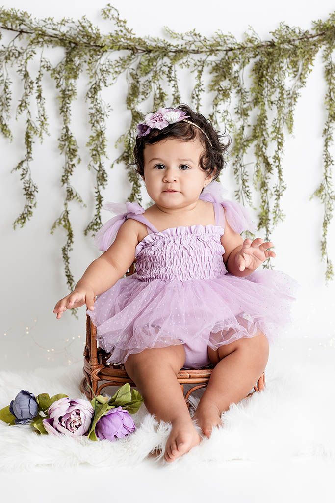 Baby girl in lavender tutu romper with floral headband sits on chair with purple flowers and greenery backdrop.