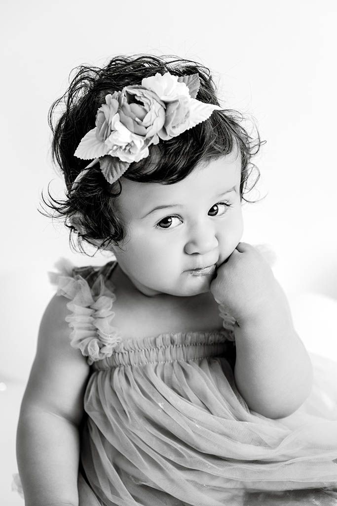 Black and white portrait of a cute baby girl in a flowy dress with a floral headband, posing thoughtfully.