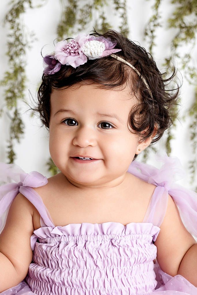 Smiling baby girl in lavender ruffle dress with floral headband, dark curly hair, portrait photo.