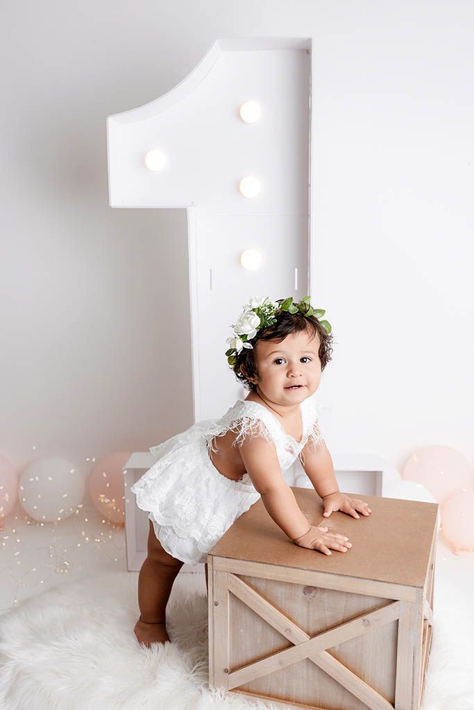 Baby girl in white lace romper and floral crown celebrates first birthday beside large number one prop.