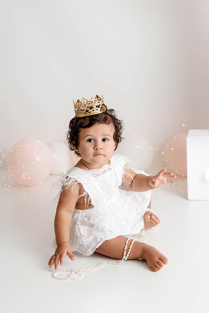 Baby girl in white lace romper and gold crown sits with pearl necklace and pink balloons for first birthday cake smash.