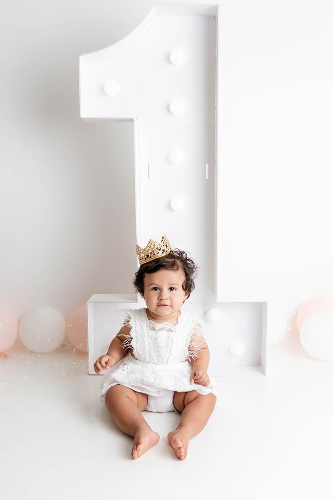 Adorable baby girl in white dress and gold crown sits in front of a large marquee number one for her first birthday.