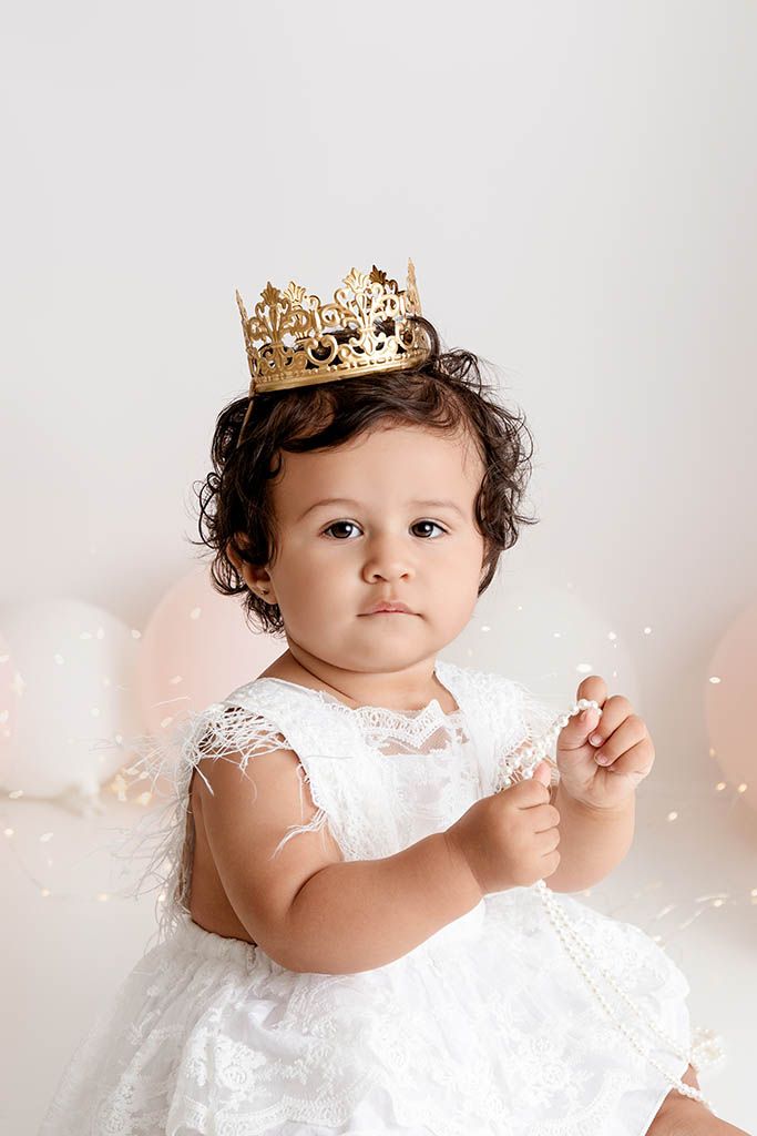 Baby girl in a white lace dress wearing a gold crown sits among balloons for her 1st birthday photo by Wildomar 1st birthday photographer.