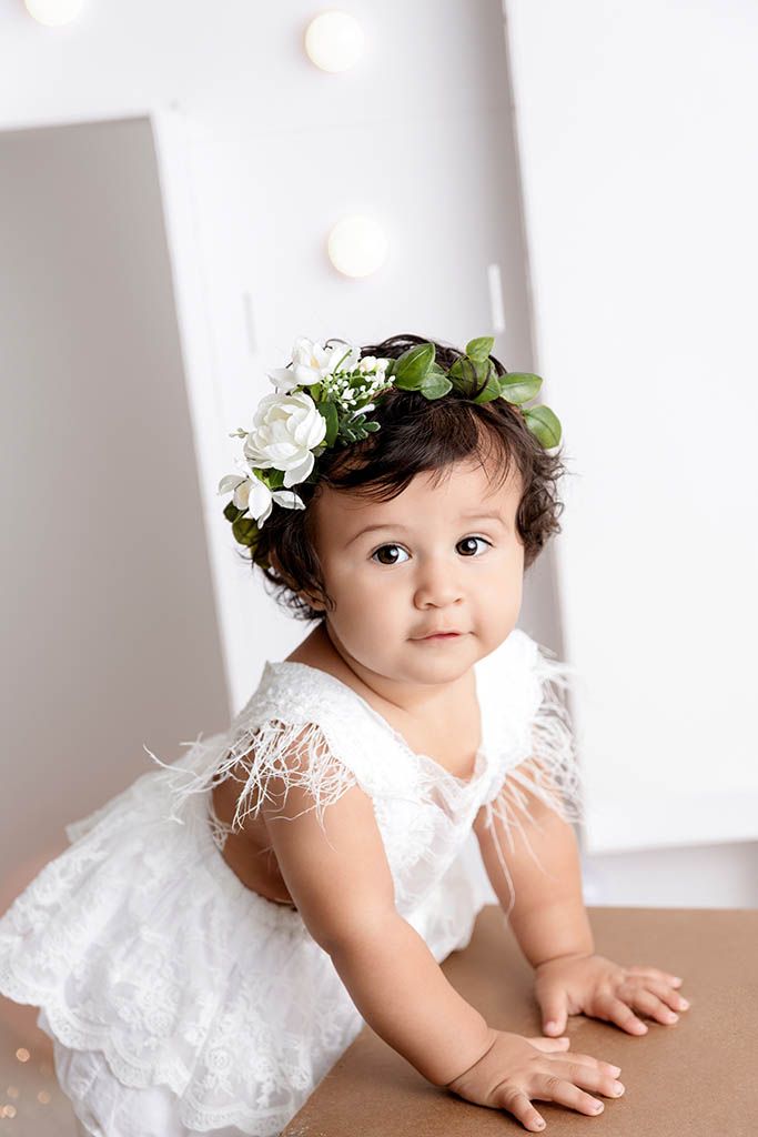 Adorable baby girl in white lace dress and floral crown with white roses and greenery, posing for a portrait photo.