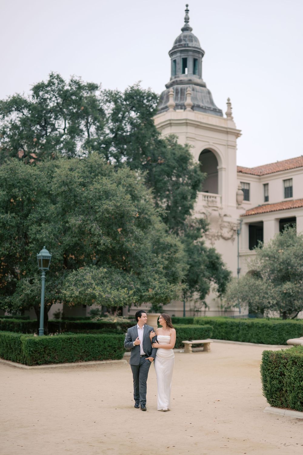 Couple walks hand in hand in formal attire before a grand historic building with a tower and lush green hedges.