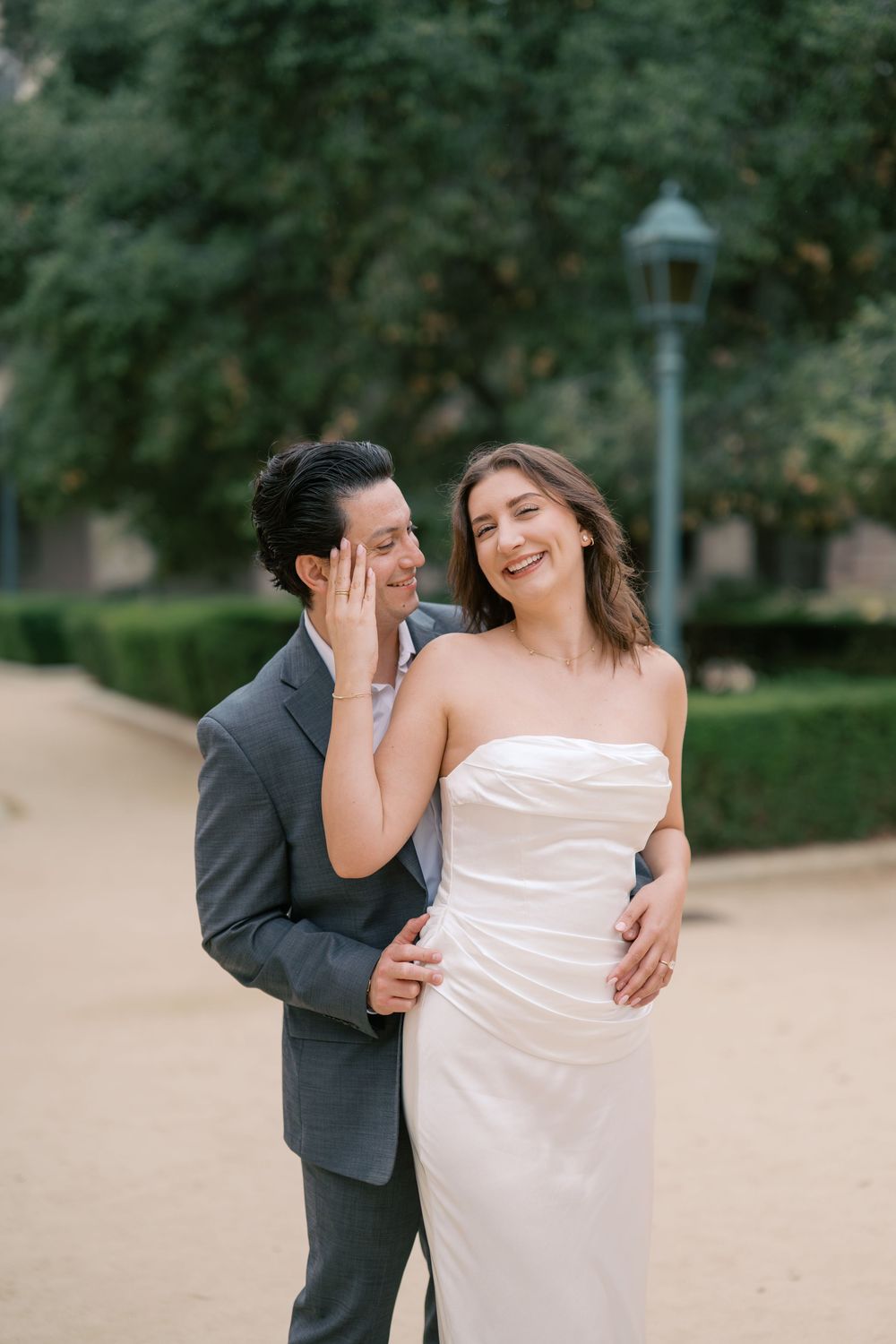 Bride and groom share a joyful moment outdoors, she in a strapless white gown, he in a gray suit.